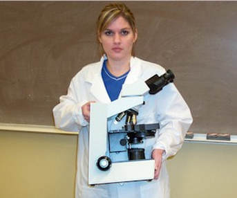 Image of a female student holding a compound microscope properly, with one hand on the base of the microscope and the other hand on the neck of the microscope. 